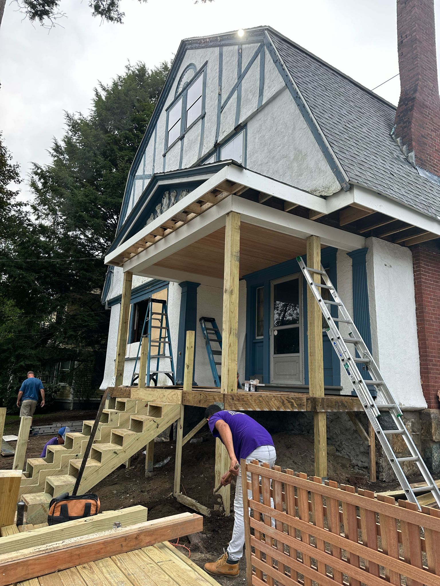 Tudor-style home addition with deck construction showing workers building custom porch with stairs